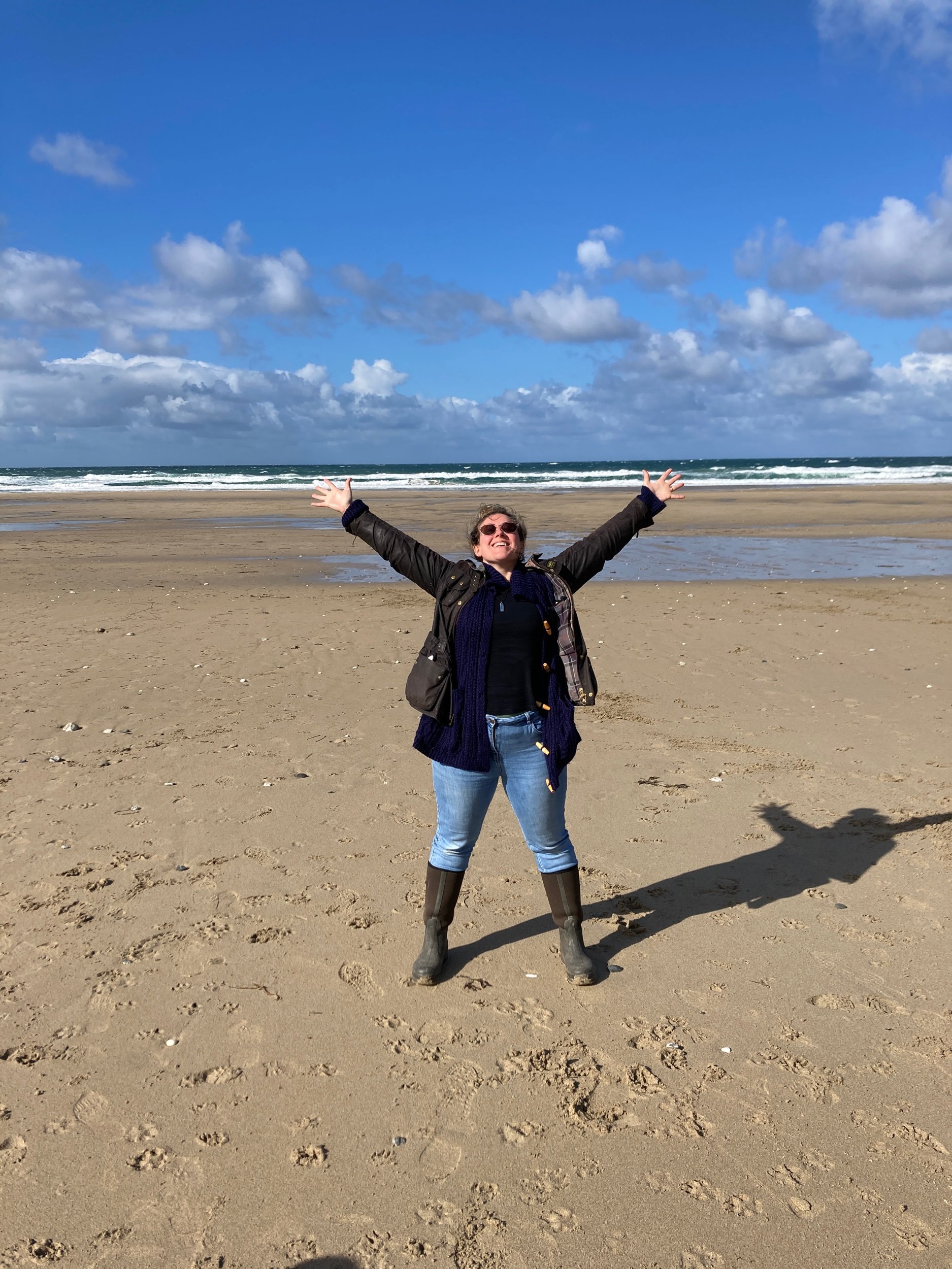 A woman stood on a beach in winter. She is in a celebratory posture, legs apart and grounded in the sand. Her arms are reaching up and out toward the blue sky. In the background the sea lines the landscape and her face apears joyful and happy.