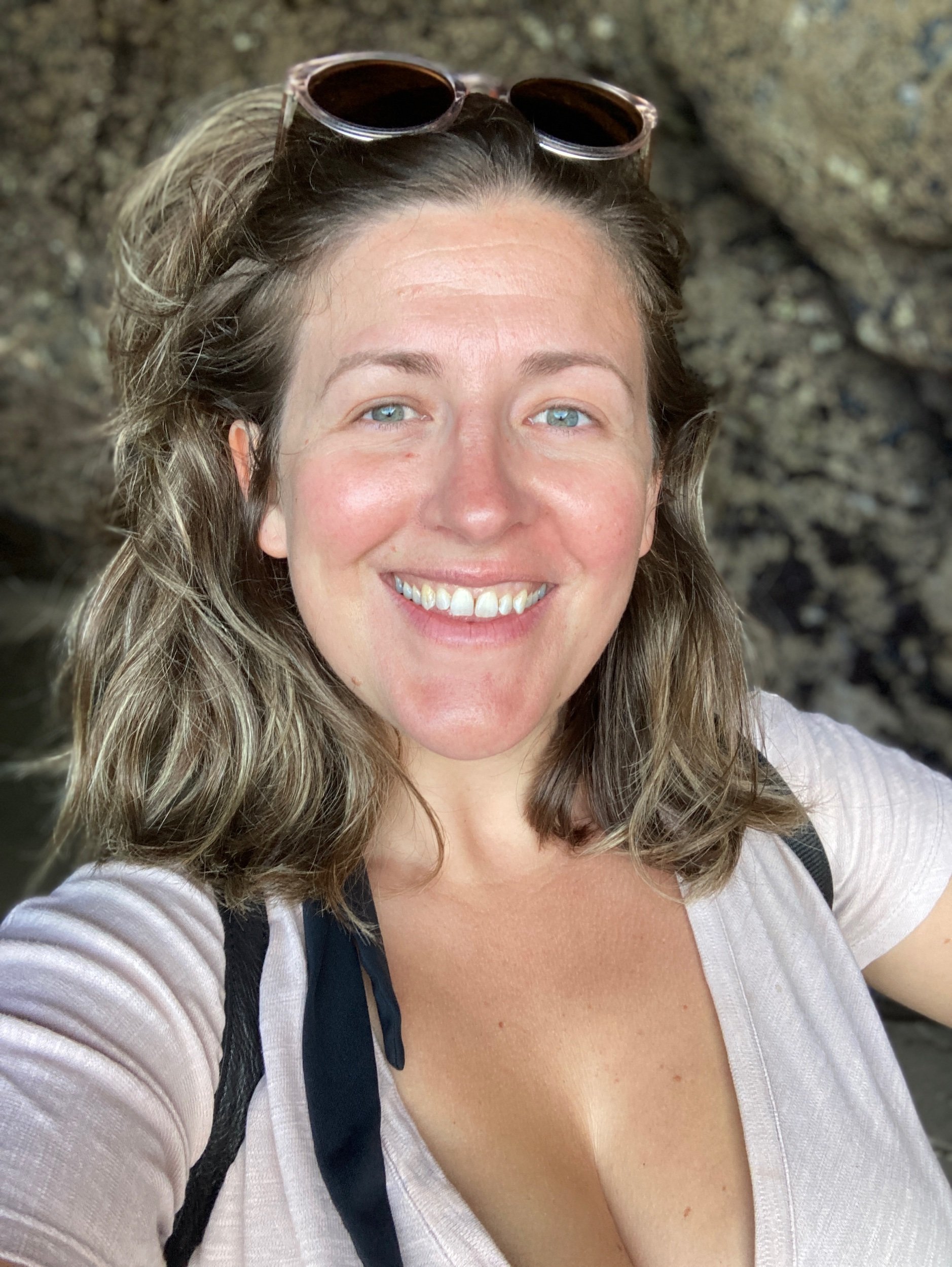A portrait selfie picture of a woman. The image is quite close up and she looks relaxed and happy. She is smiling, she appears to be wearing summer cloths, her hair is shoulder length, natural and is held back by sunglasses which she wears on top of her head. There is a cave in the background and it looks as if she has been swimming in the sea.