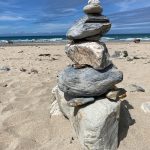 Pebbles stacked up on a beach to create a structure. The sand is soft, the sky is blue and there is an imperfection to the structure that makes it look different to a regular image of pebbles balancing.