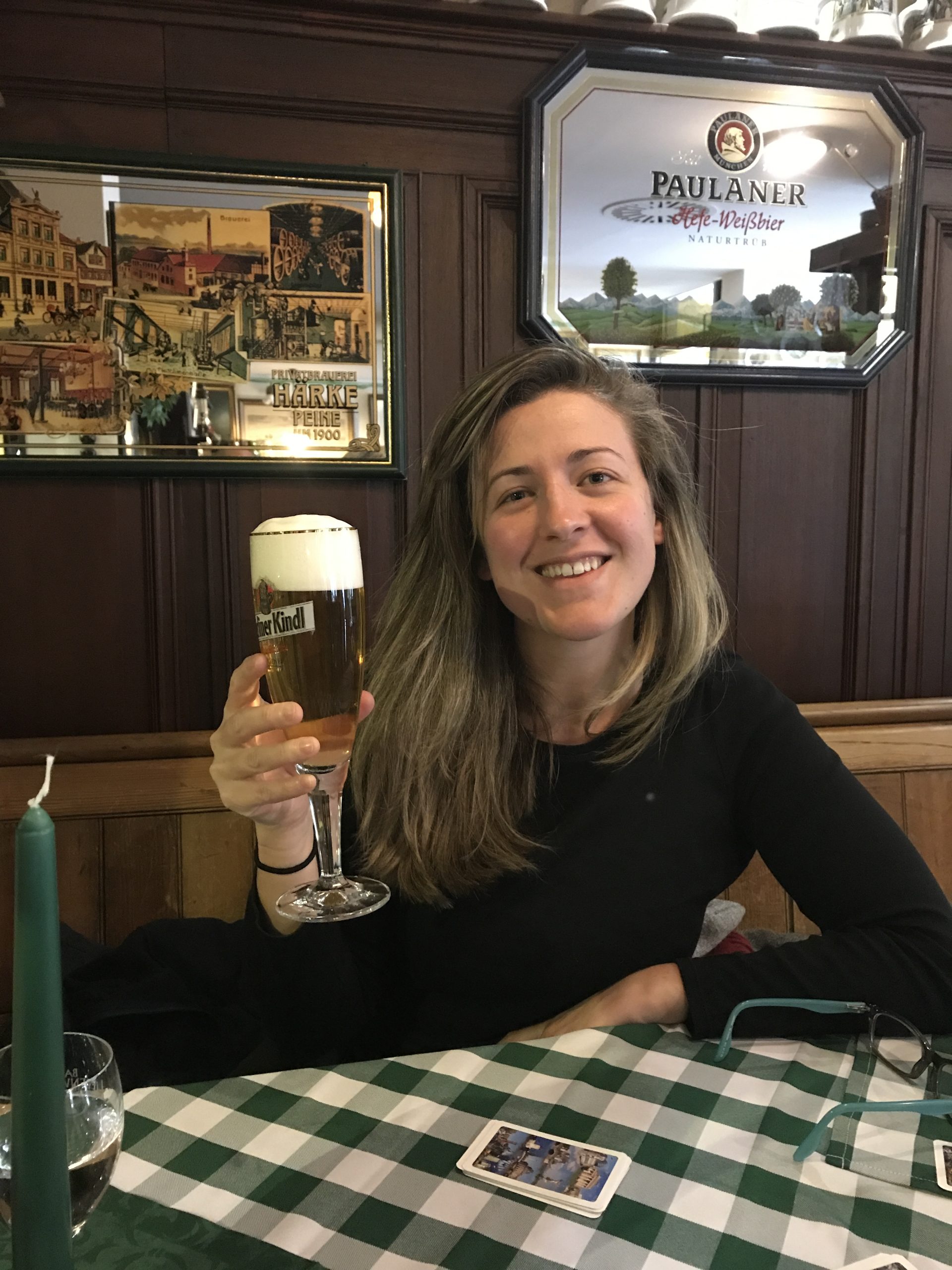 A woman sat in a bar in Germany with a been, lifting it up and smiling. She has her elbow resting on the table and she looks relaxed.