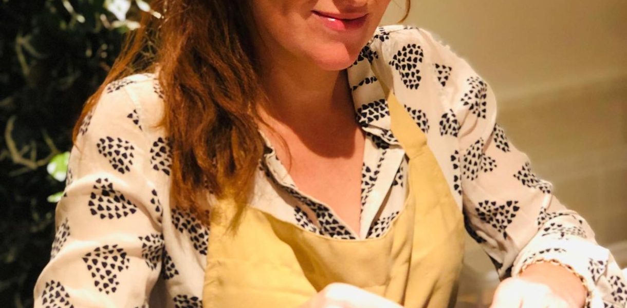 a woman with dark red hair worn in a ponytail sitting at a table blending essential oils. She is wearing a yellow apron and looking down at her work with a calm and happy expression.