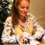 a woman with dark red hair worn in a ponytail sitting at a table blending essential oils. She is wearing a yellow apron and looking down at her work with a calm and happy expression.