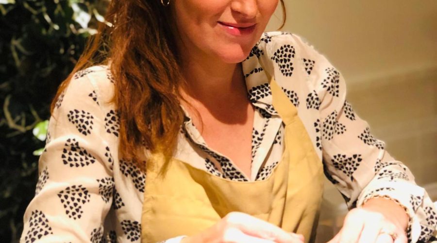 a woman with dark red hair worn in a ponytail sitting at a table blending essential oils. She is wearing a yellow apron and looking down at her work with a calm and happy expression.