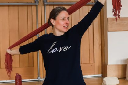 A woman sitting teaching a yoga class. She is wearing her hair tied back and holds a red scarf in her hands are she stretches her arms up and out. Her legs are crossed and she has a doll in front of her on a yoga mat. In the background a is a double door with a fire exit sign on.